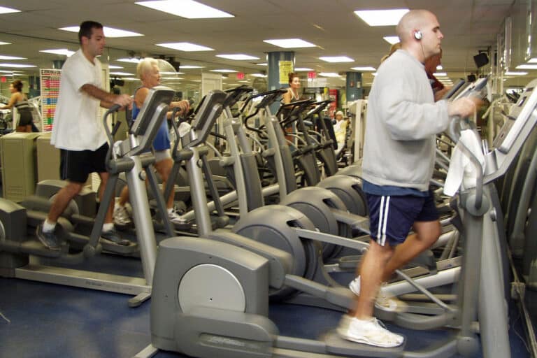 Qual a função do aparelho elíptico e quais são os seus benefícios para a saúde? 2 homens usando aparelho elíptico