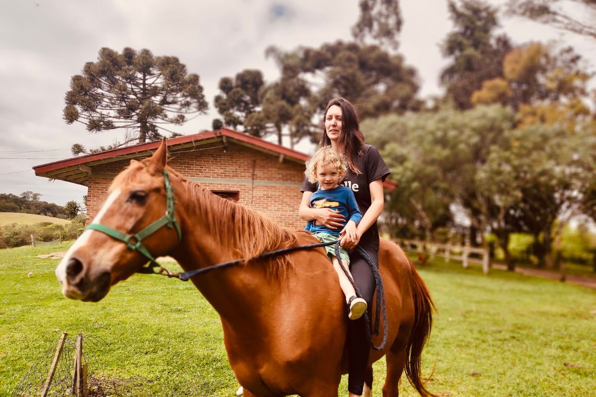 Heloísa Santos, uma mulher branca de cabelos pretos, com seu filho Oliver, uma criança pequena. Eles estão montados em um cavalo, com uma casa e árvores ao fundo.